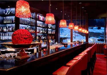 Eine stilvolle Bar mit roten Lampenschirmen und einer eleganten Einrichtung. Im Hintergrund sind schneebedeckte Landschaften zu sehen.