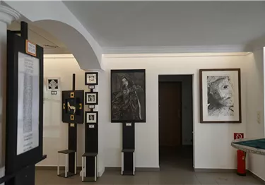 Ein moderner Raum mit mehreren Kunstwerken an den Wänden. Es gibt auch Informationsstände und Sitzgelegenheiten im Bereich.