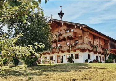 Ein traditionelles Holzhaus mit Balkonen und bunten Blumen. Im Vordergrund steht ein Baum und der Himmel ist klar.