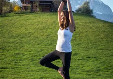 Eine Frau praktiziert Yoga in der Baum-Position auf einem Felsen vor einer Bergkulisse.